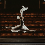 Photograph of a ballet dancer at the apex of a jump in a full split, with translucent white skirt and sleeves illuminated against the dark background of an empty theater.