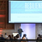John Sherry speaking on stage at EPIC2022. Projected slide says "How is remote work affecting resilience?"